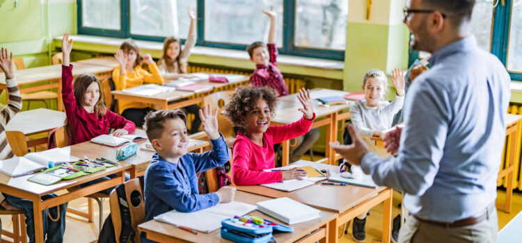 Large group of happy elementary students raising their hands to answer the teacher's question on a class in the classroom.