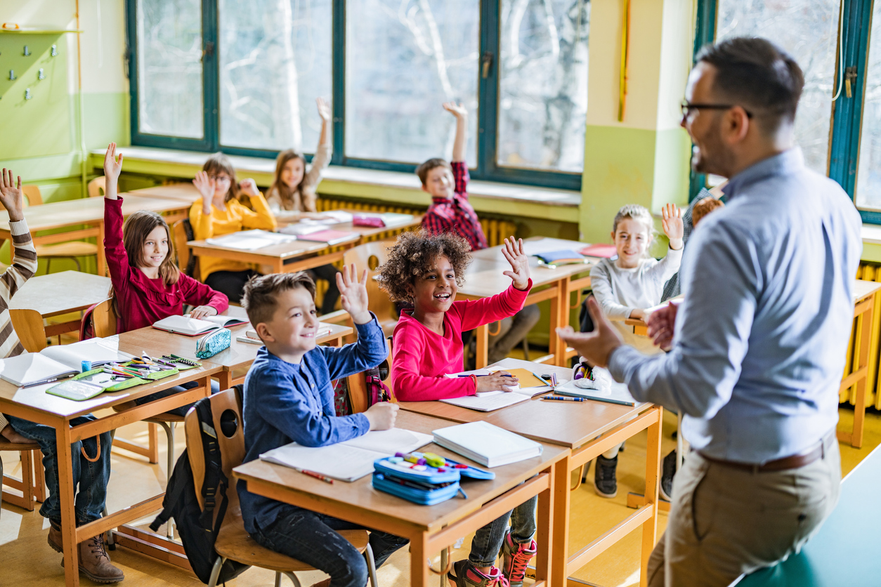 Large group of happy elementary students raising their hands to answer the teacher's question on a class in the classroom.