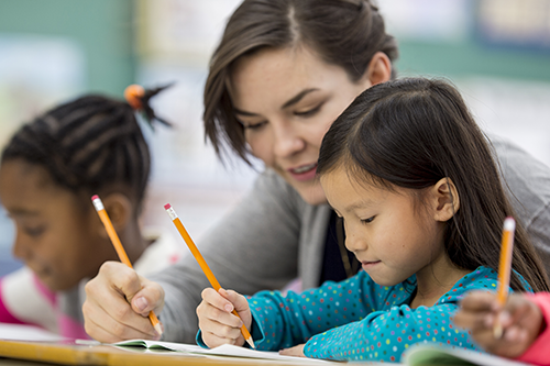 Teacher and student are writing in a workbook together, with another student in the background also working.